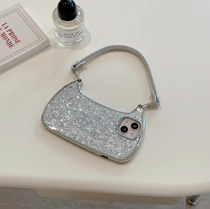 An Electroplated Handbag Hand Strap Phone Case for iPhone by curved cases, with glitter metallic waves creating a silver sparkle, sits atop a white surface. Nearby is a rectangular book titled "LA PROSE DU MONDE" with red text and a clear bottle of perfume labeled "ROSE NOIRE." The handbag's glitter matches the phone case inside perfectly.