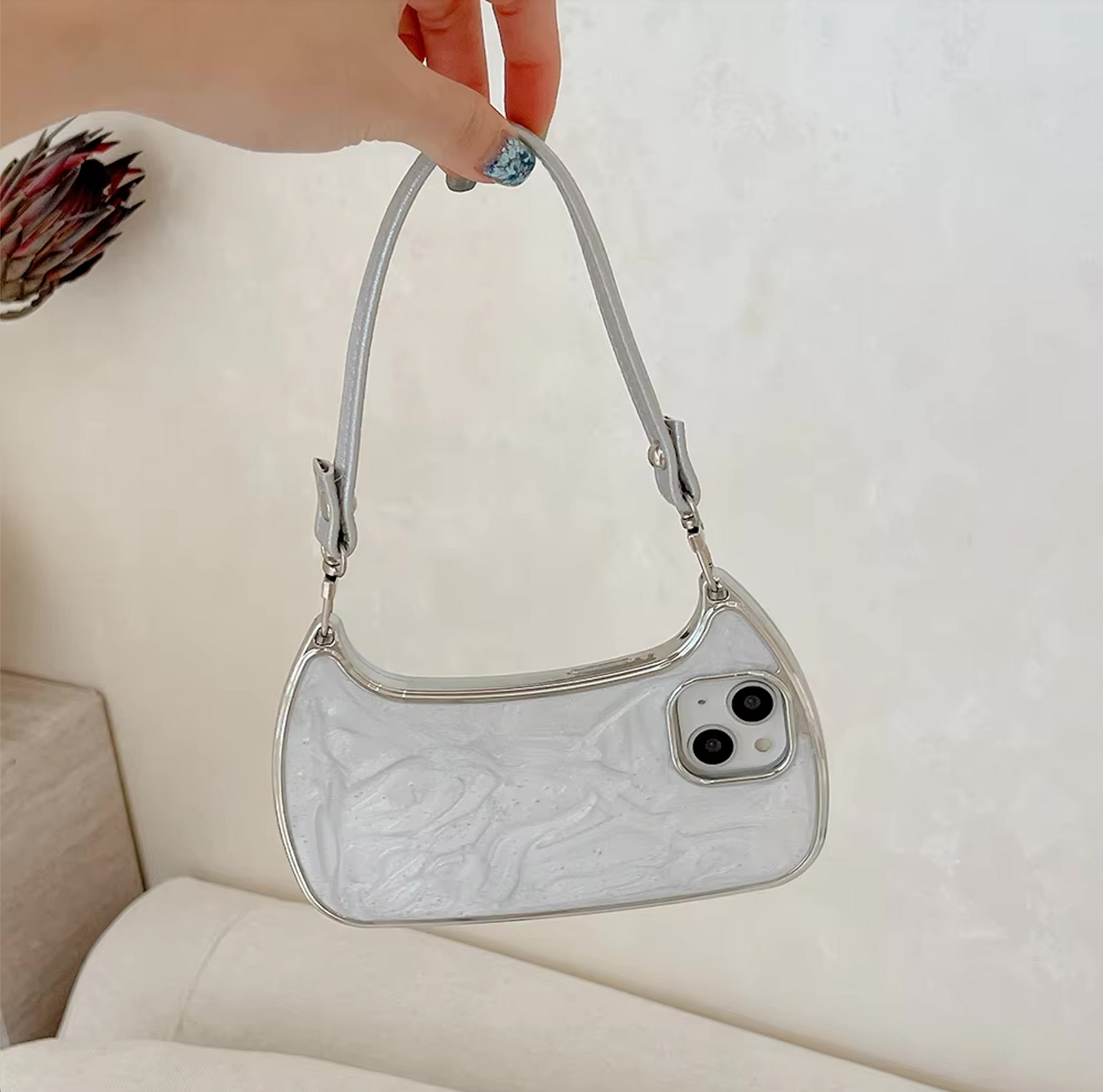 A hand holds the Electroplated Handbag Hand Strap Phone Case by curved cases. This small, shiny white accessory features a silver zipper and short handle, resembling a handbag but designed as a phone case for iPhone models 15 to 7 Plus. The distinctive design includes an imitation phone camera lens on one side. In the background, you can see an abstract wall and part of a sofa adorned with red and brown floral patterns.