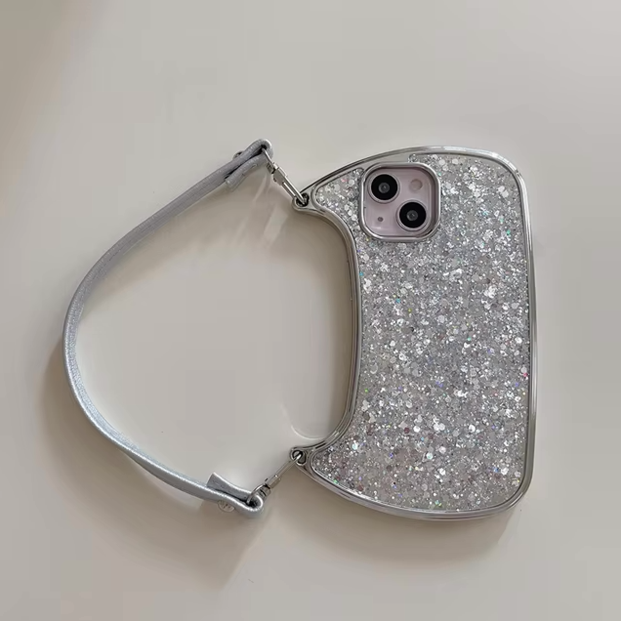 An Electroplated Handbag Hand Strap Phone Case by curved cases is lying on a white surface. This silver, glittery case is designed to fit an iPhone with a dual-camera setup and features an anti-scratch finish. The attached matching handle is made of a silver strap.